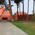 Eduardo Souto de Moura's House of Stories Paula Rego showcasing a striking red concrete structure with composite materials, surrounded by a shaded lawn and a concrete walkway.