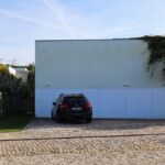 Nuno Graça Moura's 2 Villas at Bom Sucesso Resort showcasing a modern design with a concrete driveway leading to a garage door, accompanied by a parked subcompact car and sedan.