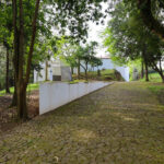 Álvaro Siza Vieira’s Piscina da Quinta da Conceição showcasing a minimalist concrete structure integrated with landscaped garden elements, shaded walkways, and surrounding woody plants including branches and trunks under natural sunlight.