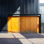 Casa Sacerdotal de São Martinho de Dume by André and António Fontes showcasing a modern structure with wood and composite materials, featuring stained hardwood planks, metal accents, and detailed wood flooring under natural shadow.