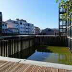 Casa Sacerdotal de São Martinho de Dume by André and António Fontes showcasing a high-rise condominium with composite material façade, balconies, and reflective glass panels overlooking an urban canal during daytime.