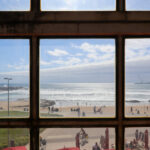 Carlos Prata and Manuel de Solà-Morales’ Edificio Transparente showcasing a modern glass façade with cantilevered balconies overlooking a sunlit coastal bay with sandy beach and ocean shore.