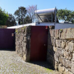 Eduardo Souto de Moura’s Casa 1 em Miramar showcasing a stone and brick wall façade with concrete elements, a driveway leading to a gated entrance, and a walkway bordered by textured building materials.