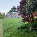 Eduardo Souto de Moura’s Casa 1 no Bom Jesus showcasing a minimalist modern design with clean lines, surrounded by landscaped garden featuring grass, shrubs, groundcover, and hedges.