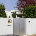 Eduardo Souto de Moura’s Casa Rua da Índia showcasing a concrete gate with composite material accents, a shaded driveway, home fencing, and a neatly trimmed hedge.