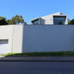 Eduardo Souto de Moura’s Casa do Cinema Manoel de Oliveira showcasing a minimalist concrete and plaster façade with composite material elements, featuring shaded areas and daylighting effects over a driveway with distinct shadows.