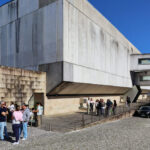 Jorge Nuno Monteiro’s Pavilhão Desportivo Municipal de Santo Tirso showcasing brutalist architecture with concrete walls, composite materials, and daylighting through large openings, featuring prominent stairs and textured surfaces.