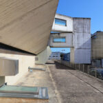 Jorge Nuno Monteiro’s Pavilhão Desportivo Municipal de Santo Tirso showcasing a daytime view of the concrete and metal composite structure with urban design elements and adjacent commercial and residential buildings.