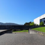 OPERA | Design Matters' Escola de Medicina, Universidade do Minho, showcasing a modern building with a sleek glass façade and angular structural elements under a sky with cumulus clouds.