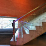 Rem Koolhaas’s Casa da Música interior showcasing composite material flooring, wooden stained stairs with metal handrails, and large glass panels enhancing the modern architectural design.