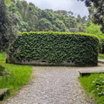 Unknown Architect’s Shower House Parque da Cidade showcasing a stone wall structure integrated with surrounding shrubs, hedges, groundcover, and landscaped garden pathways providing shaded environmental art.