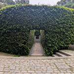 Unknown Architect’s Shower House Parque da Cidade showcasing a modern structure surrounded by shrubs, hedges, and a botanical garden with shaded walkways and evergreen plants.