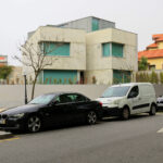Unknown Architect’s Villa I Praça de Liége showcasing a modern residential design with a sleek glass façade and integrated green rooftop, visible alongside a parked family car with illuminated automotive tail and brake lights.