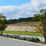 Nuno Graça Moura’s Golf Clubhouse at Bom Sucesso Resort showcasing a landscaped garden with stone walls, shrub and groundcover plantings, a botanical garden area, and a winding walkway path.