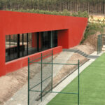 Nuno Graça Moura’s Tennis Club at Bom Sucesso Resort showcasing a red-shaded sports venue with artificial turf, landscaped lawn, mesh fencing, and home fencing elements.