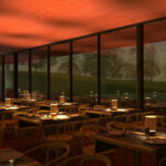 Nuno Graça Moura’s Tennis Club at Bom Sucesso Resort showcasing a wood-stained restaurant interior with hardwood floors, varnished dining tables, chairs, and a kitchen area illuminated at dusk.