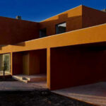 Atelier dos Remédios, Madalena Cardoso de Menezes, and Francisco Teixeira Bastos' 18 Red Row Houses at Bom Sucesso Resort featuring composite and concrete materials with orange accents, showcasing sharp shadows and daylighting effects in the evening.