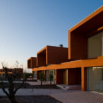 Atelier dos Remédios, Madalena Cardoso de Menezes and Francisco Teixeira Bastos’ 18 Red Row Houses at Bom Sucesso Resort showcasing a composite material façade with modern building materials illuminated in the evening.