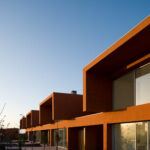 Atelier dos Remédios, Madalena Cardoso de Menezes, and Francisco Teixeira Bastos’ 18 Red Row Houses at Bom Sucesso Resort showcasing a modern design with red façades and linear row house structures illuminated in the evening.