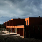 Atelier dos Remédios, Madalena Cardoso de Menezes, and Francisco Teixeira Bastos’ 18 Red Row Houses at Bom Sucesso Resort featuring a series of modern red façades with clean lines and large windows, captured at dusk.