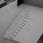 Josep Llinás’s House Model 2 showcasing a modern design with silver and plastic materials, featuring sharp shadows and paper elements in the composition.