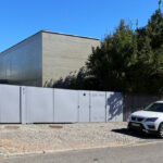Gonçalo Byrne’s Utopia Villa 3 showcasing a modern composite and concrete structure with a shaded entrance, gated driveway, garage door, and landscaped home fencing.