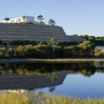 Alcino Soutinho’s 28 Unfinished Architectural Townhouses, Lot 288, showcasing a series of modern townhouses with minimalist geometric forms set against a panoramic blue sky and natural wetland landscape near a reservoir and floodplain.
