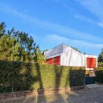 Alcino Soutinho's Habitação unifamiliar em Ofir showcasing a modern design with a landscaped garden, shaded yard, and a driveway surrounded by evergreen shrubs and hedges.