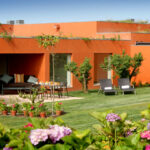 Atelier dos Remédios, Madalena Cardoso de Menezes and Francisco Teixeira Bastos’ 18 Red Architectural Townhouses, Lot 215, showcasing vibrant orange outdoor furniture including chairs and a daybed arranged in a garden backyard with houseplants and a coffee table.